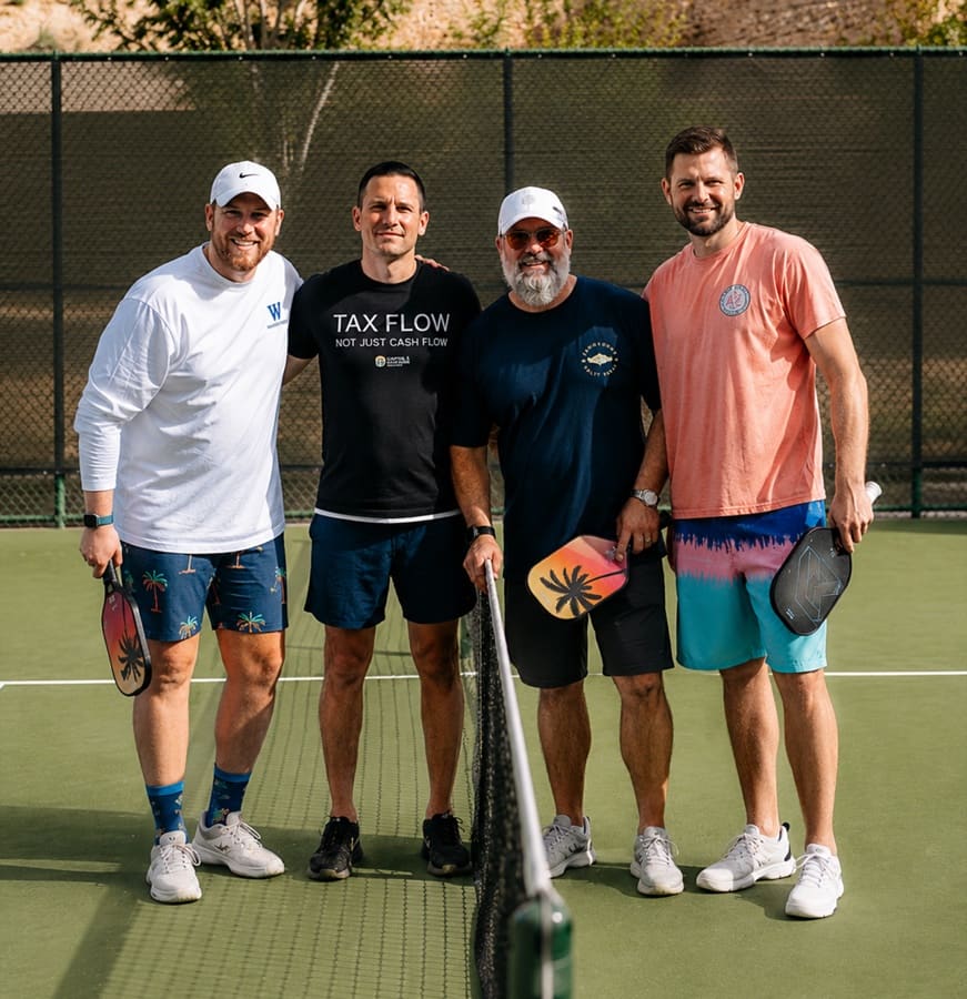 Four friends standing at the net on a pickleball court, holding paddles